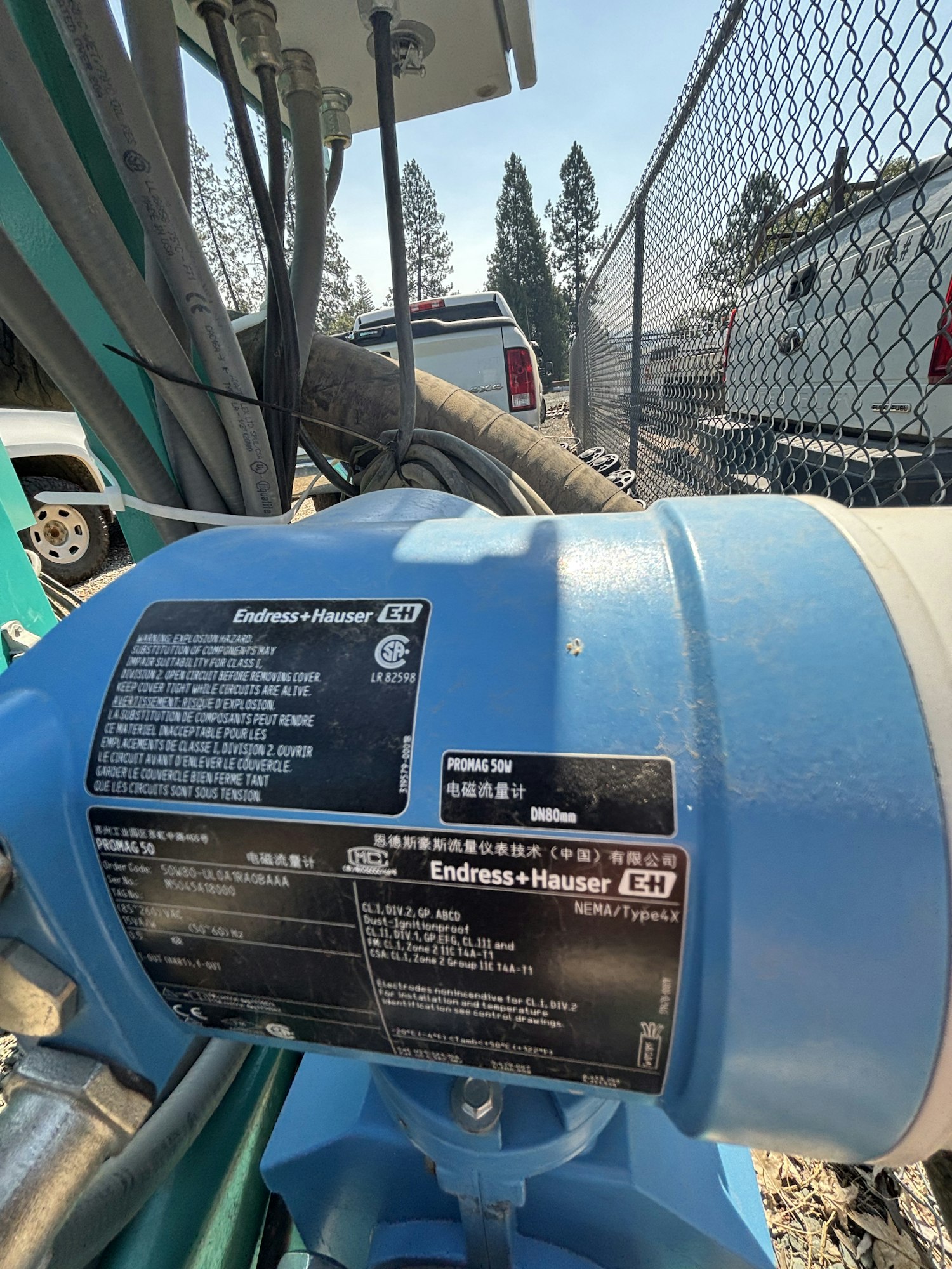 The image shows a blue Endress+Hauser PROMAG 50 flowmeter with technical specifications and wires, near parked vehicles and a fence.
