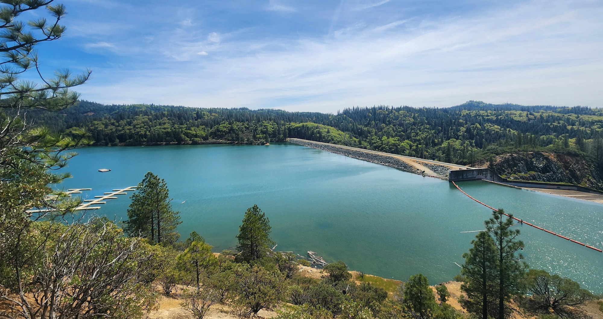 A serene lake with a dam surrounded by forested hills under a clear blue sky.