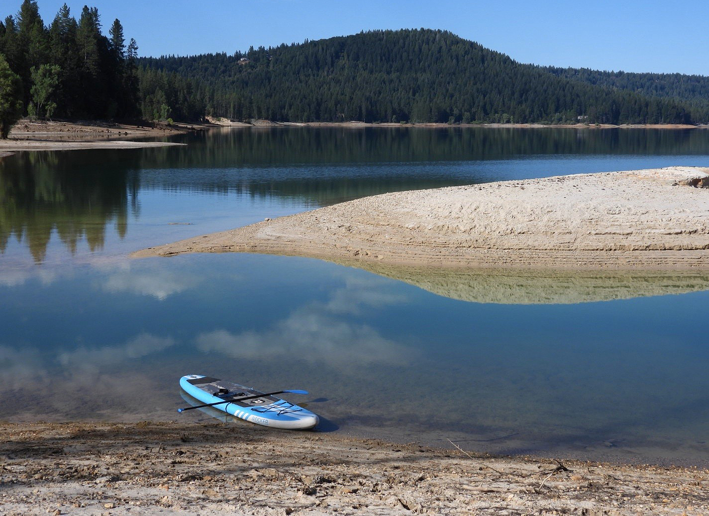 A paddleboard on the shore of a calm lake with surrounding forested hills and a clear blue sky.