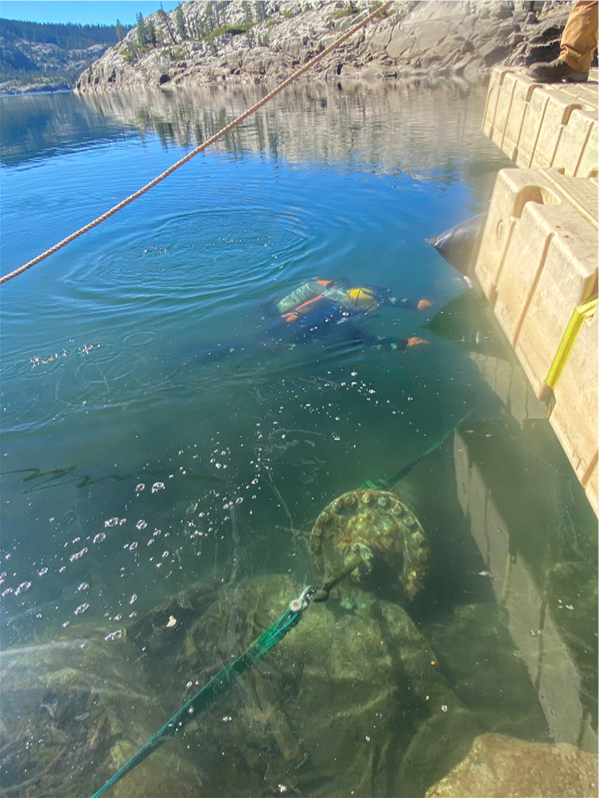 A person in a wetsuit is submerged in clear water, near a large object tethered to the shore, surrounded by rocky terrain.
