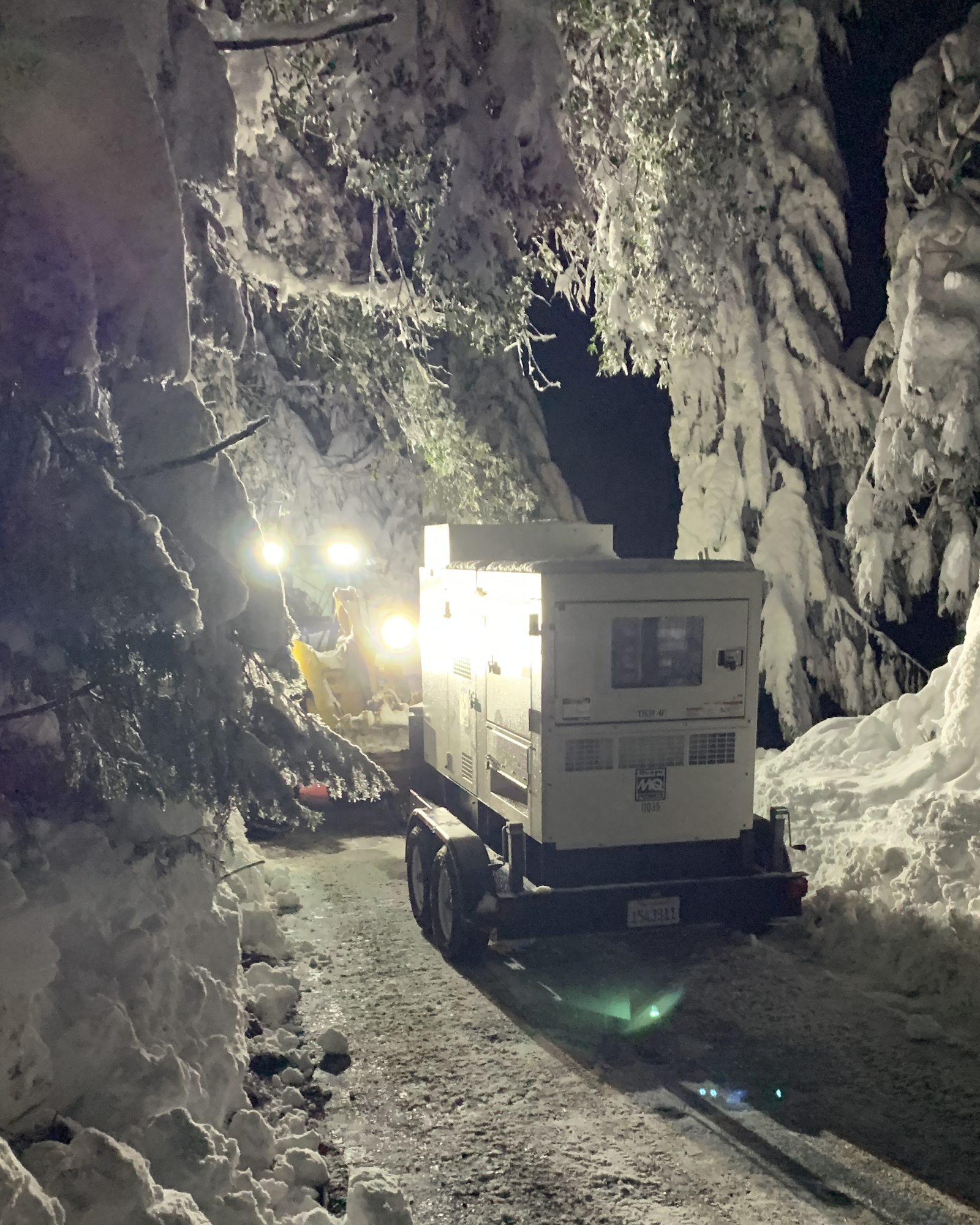 A snowy scene at night featuring a generator and bright work lights, surrounded by snow-covered trees.