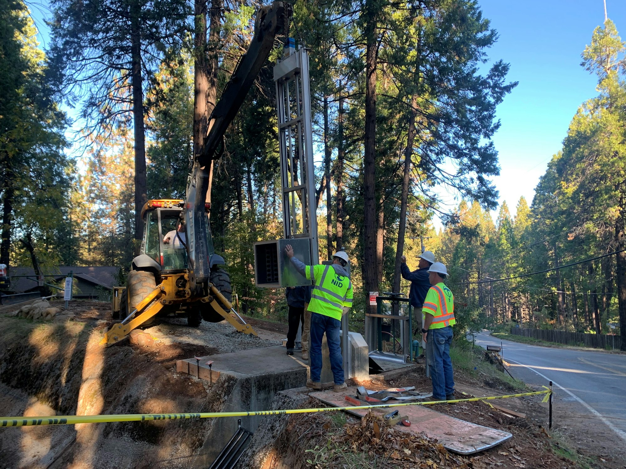 Workers are installing equipment near a road in a wooded area, using machinery for the installation process.