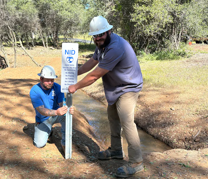 Two workers in hard hats are installing a safety sign by a canal, outlining safety rules like no swimming and no fishing.