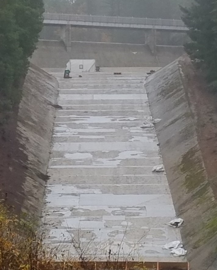 The image shows a concrete channel or dam structure with some water and debris, surrounded by trees and a bridge overhead.