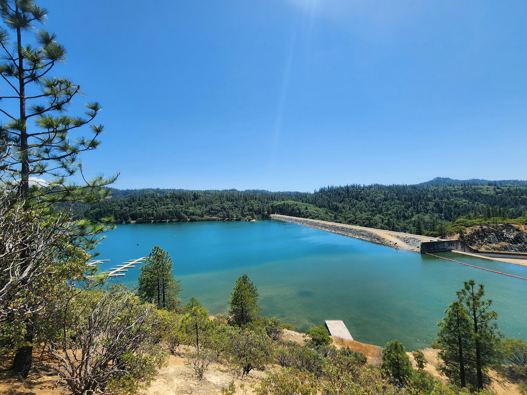 A scenic view of a lake with a dam, surrounded by forest and a clear blue sky.