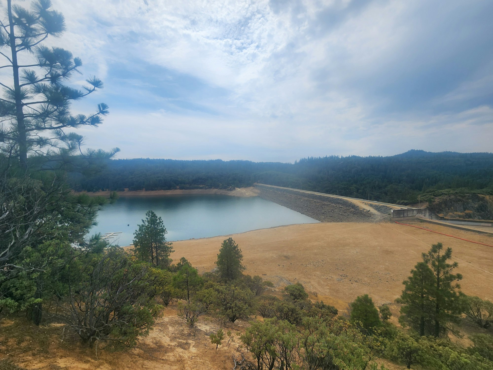 A reservoir surrounded by dry grass and forests under a cloudy sky. There's a dam structure to the right.