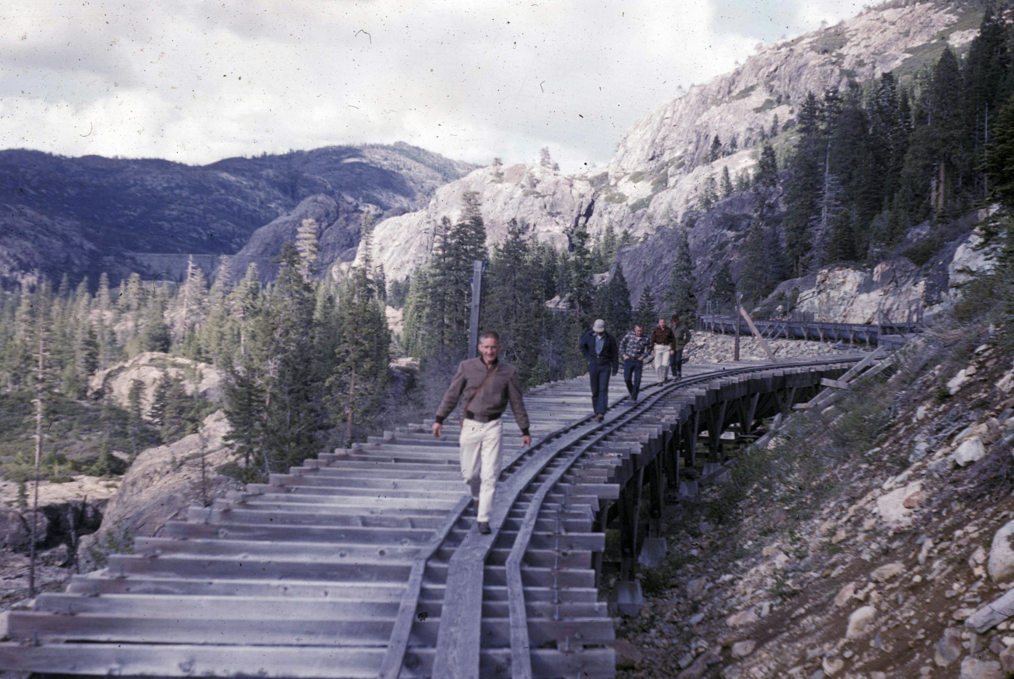A group of people walks along a wooden train track surrounded by mountains and trees in a scenic landscape.