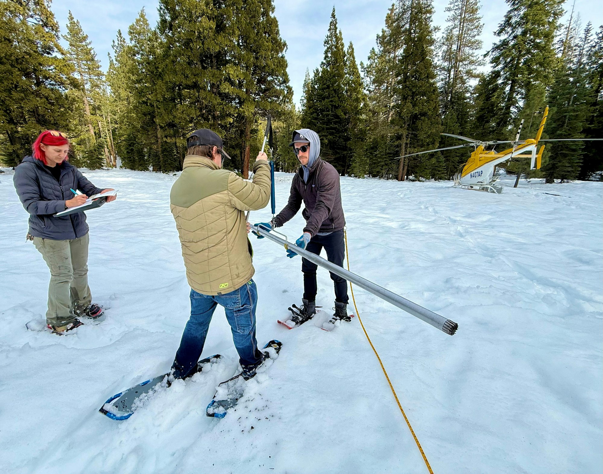 A group in snow-covered terrain conducts research, with one person taking notes and others handling equipment near a helicopter.