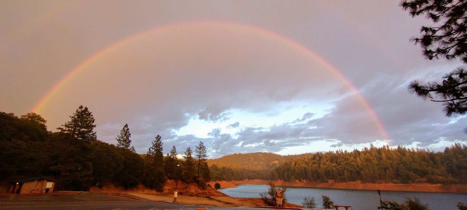 A vibrant rainbow arcs over a forest-lined lake under a pink-tinged sky, with a small building in the foreground.