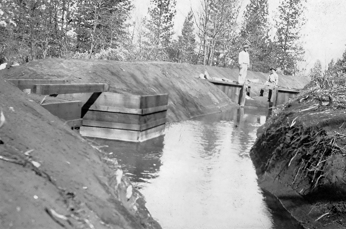 A water channel with wooden structures, two people standing on the bank, surrounded by trees.