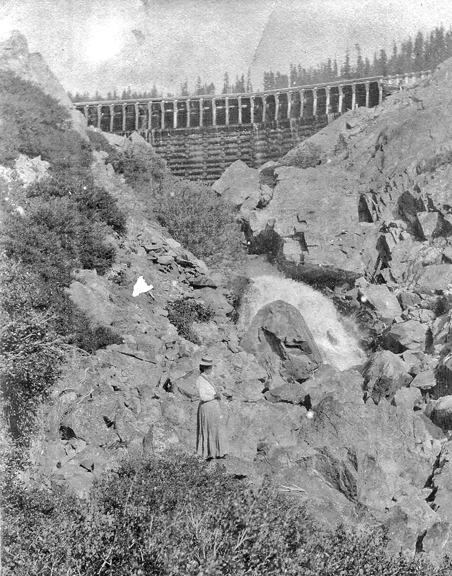A woman stands on rocky terrain near a waterfall with a large bridge in the background, surrounded by trees.