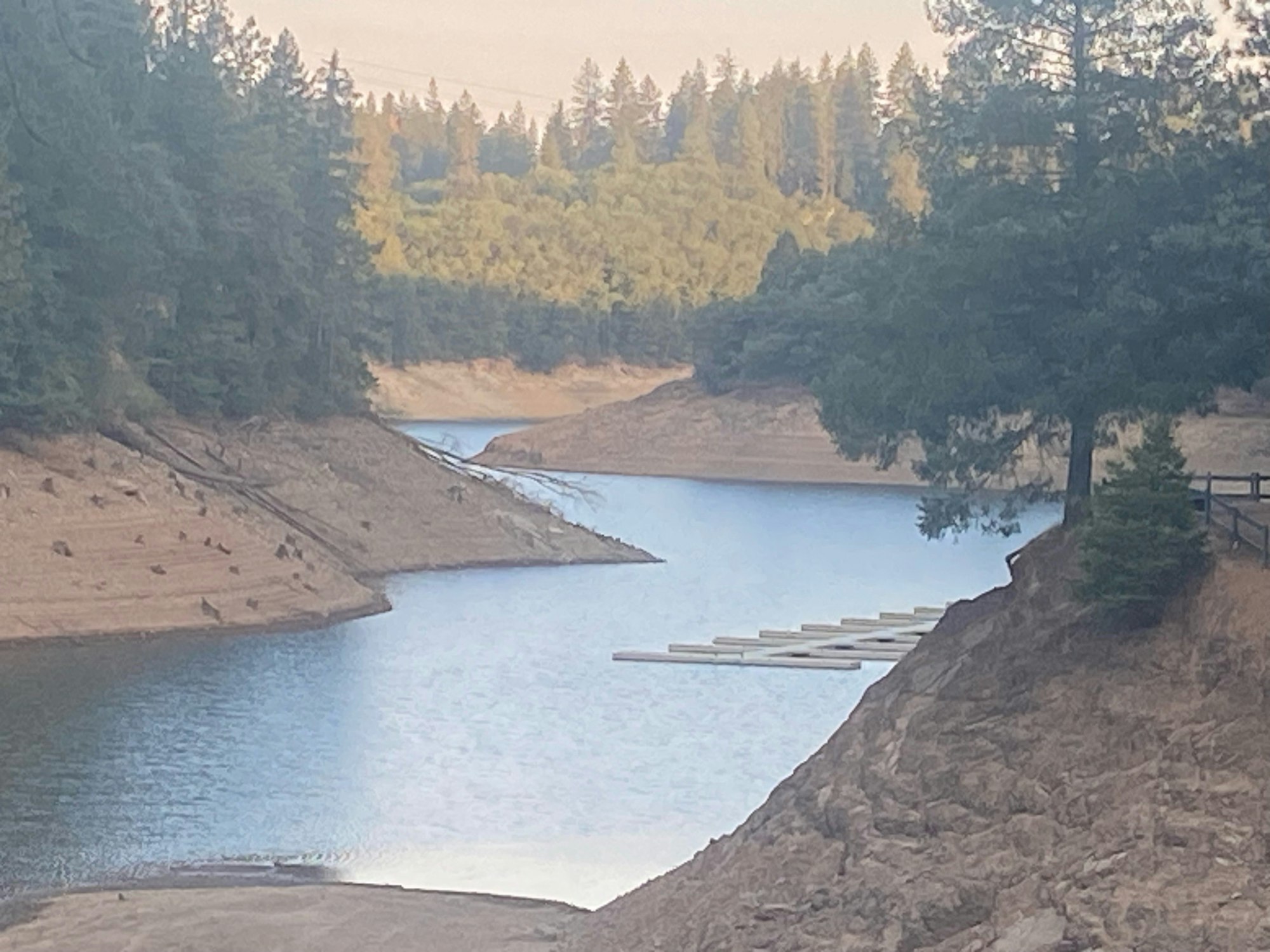 A tranquil lake surrounded by forested hills with a small dock and low water levels.