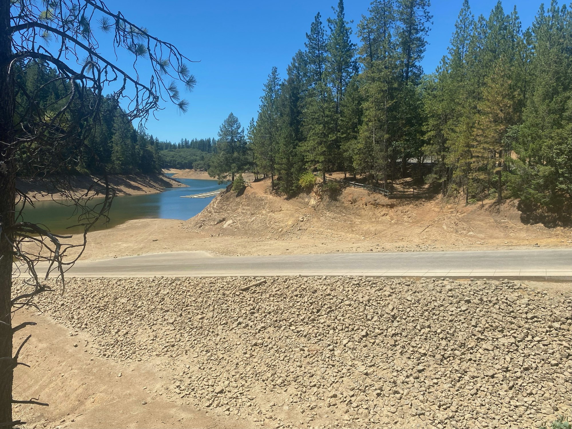 A road with a rocky shore leading to a forested hillside beside a river or lake under a clear sky.