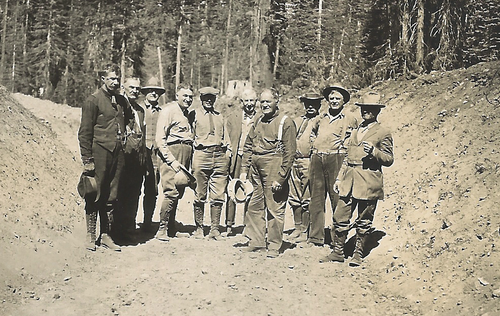 A group of men in outdoors attire standing on a dirt path in a forested area.
