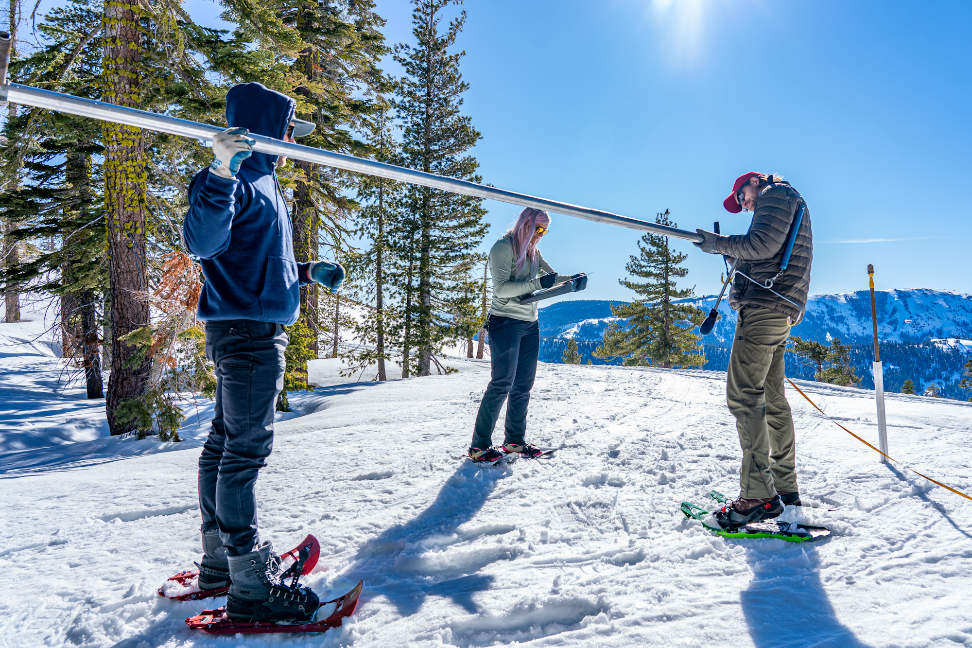 Three people in winter gear on snowy terrain, measuring with a long pole, surrounded by trees and mountains.