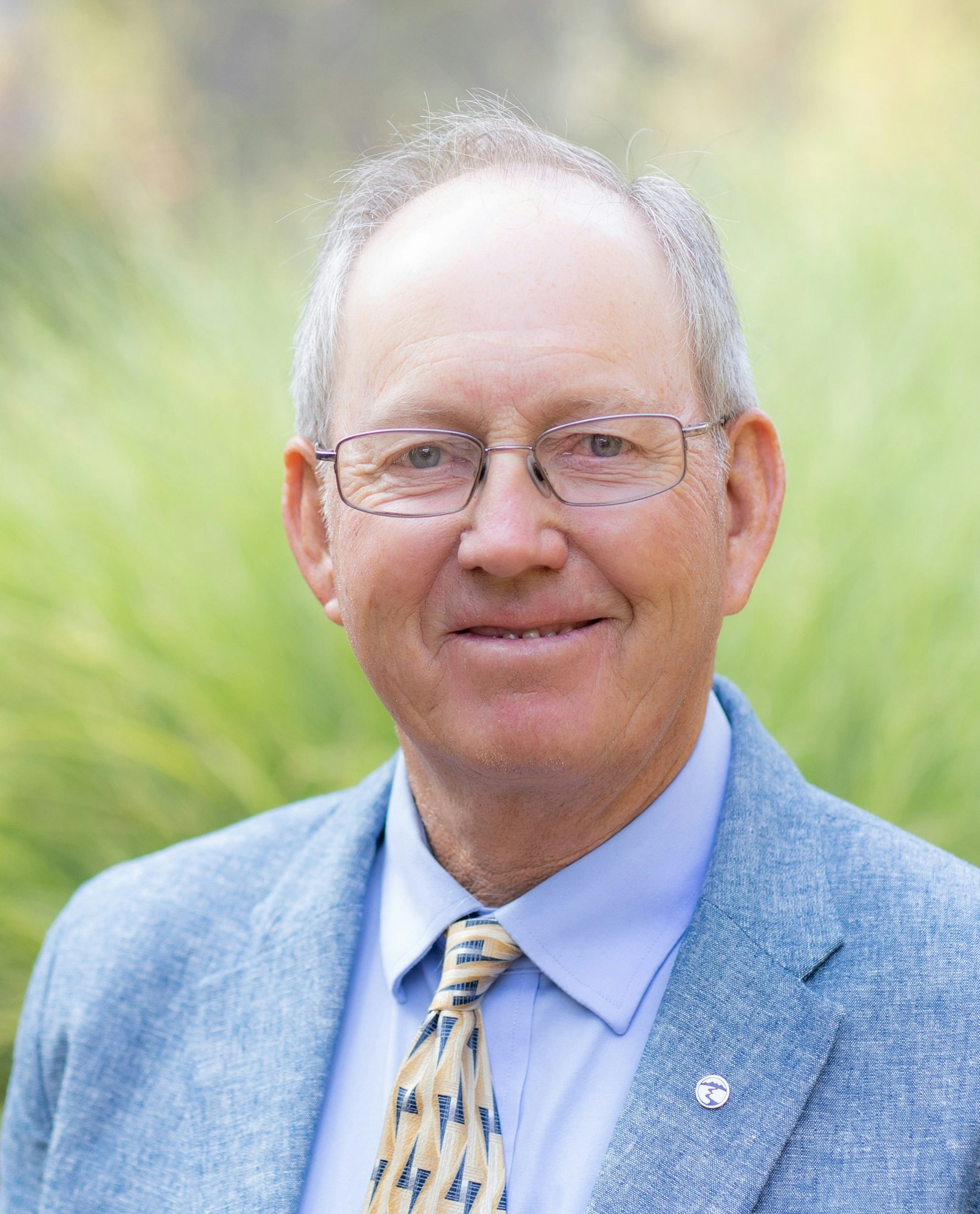 An older man wearing glasses, a light blue suit, patterned tie, and smiling, set against a blurred green background.