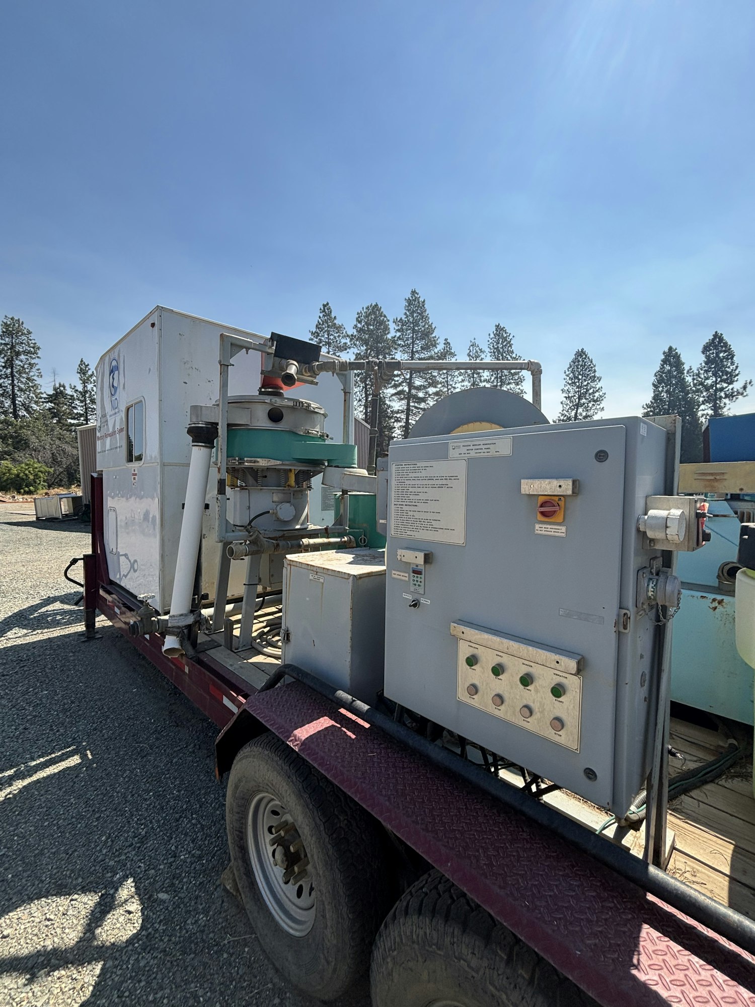 The image shows a trailer-mounted industrial machine with various controls and equipment, set against a clear blue sky and pine trees.