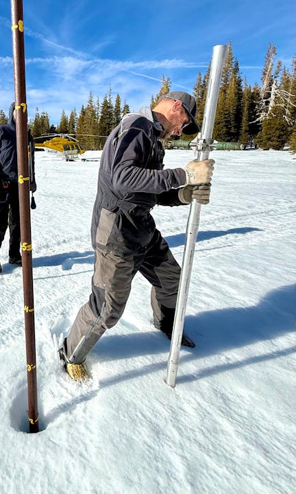 A person is using a tool to collect data from snow, with a helicopter in the background and trees around.