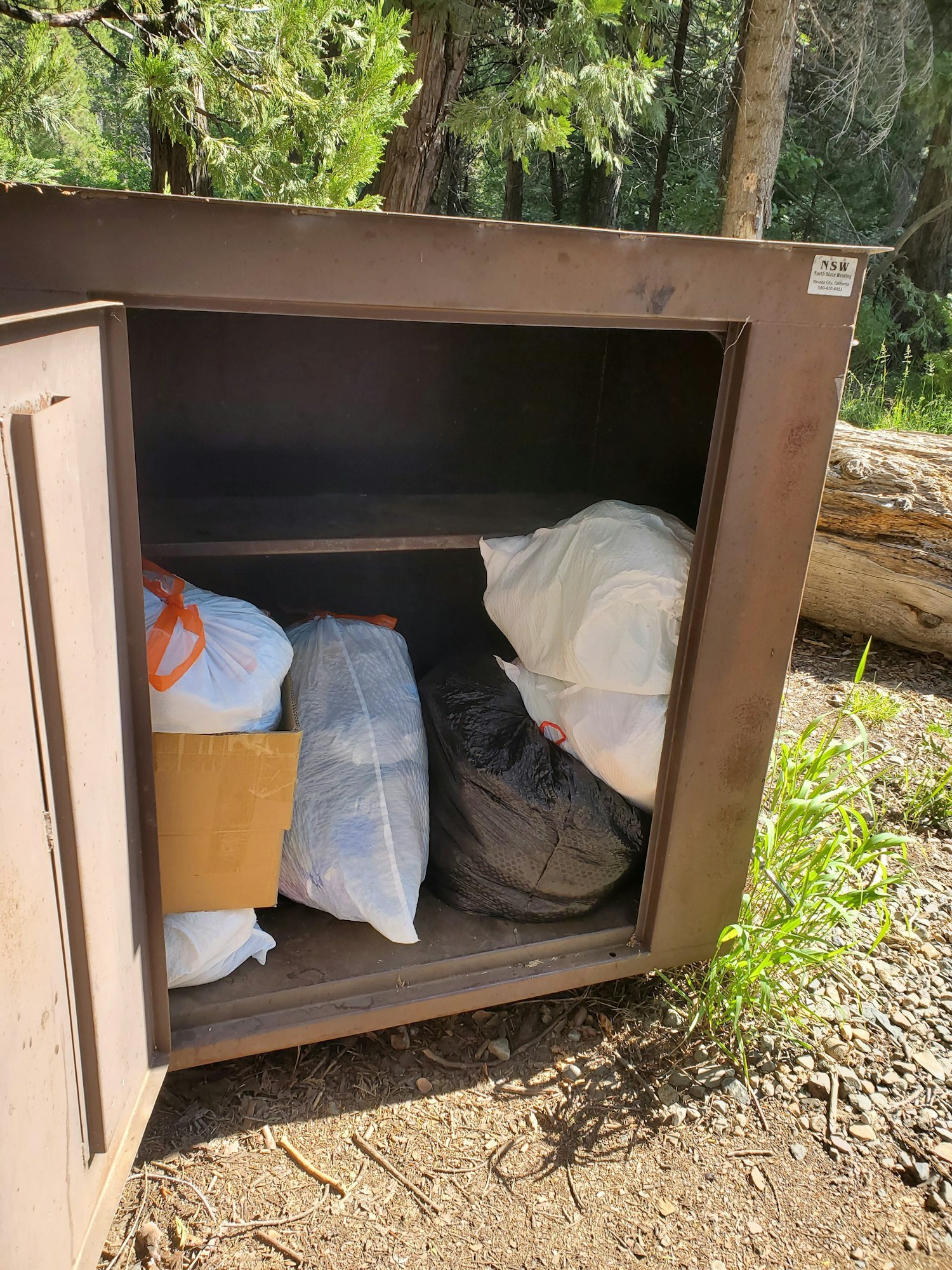 A bear-resistant garbage storage container with bags and a box inside, located in a wooded area.