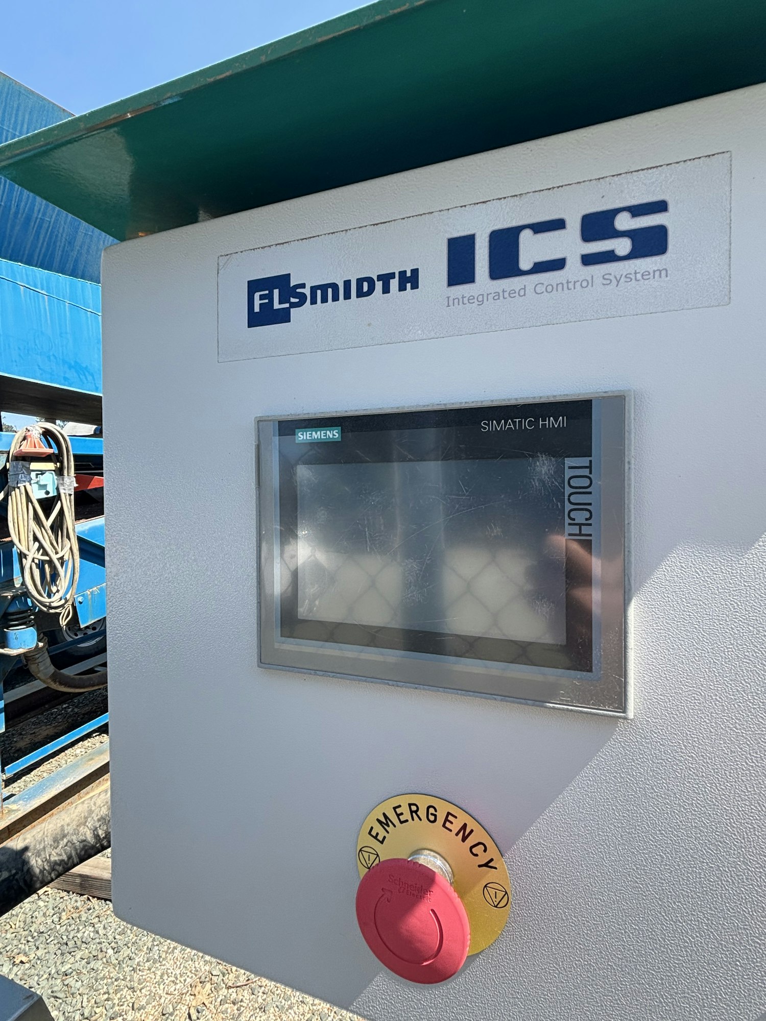 The image shows an industrial control panel with an HMI touchscreen, featuring an emergency stop button and branding from FLSmidth and Siemens.