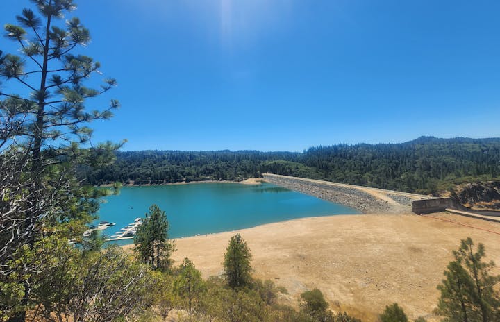 Scenic view of a tranquil blue lake, dam, marina with boats, surrounded by forested hills and a clear sky.