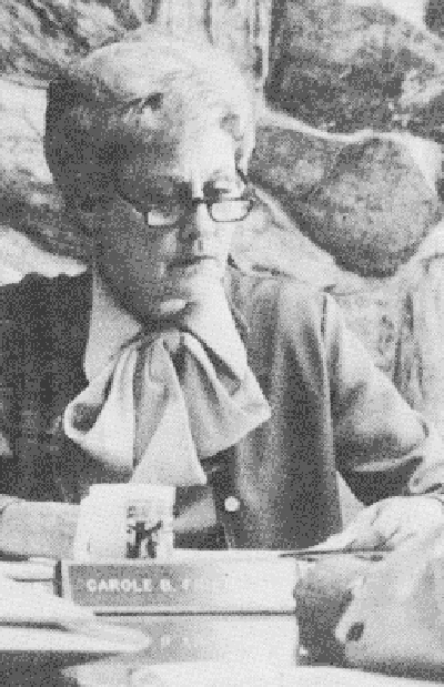 A person with glasses sitting, with a book titled "CAROLE R." and a pen in hand. The image is monochrome and grainy.