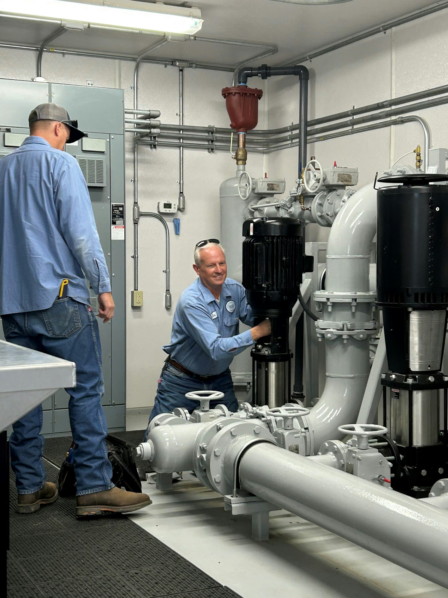 Two workers in blue shirts are servicing equipment in a mechanical room with large pipes and machinery.