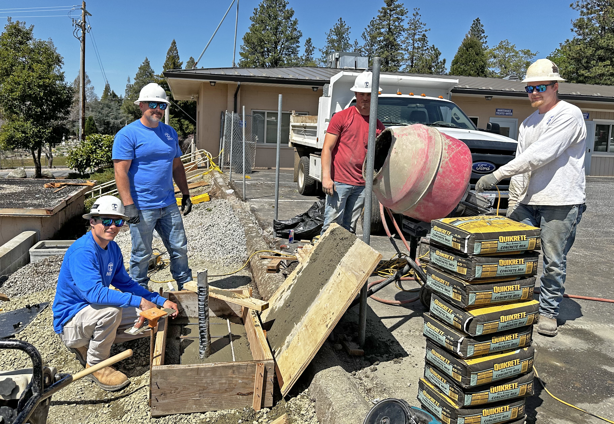 Four construction workers mixing and pouring concrete at a worksite, surrounded by Quikrete bags and equipment.