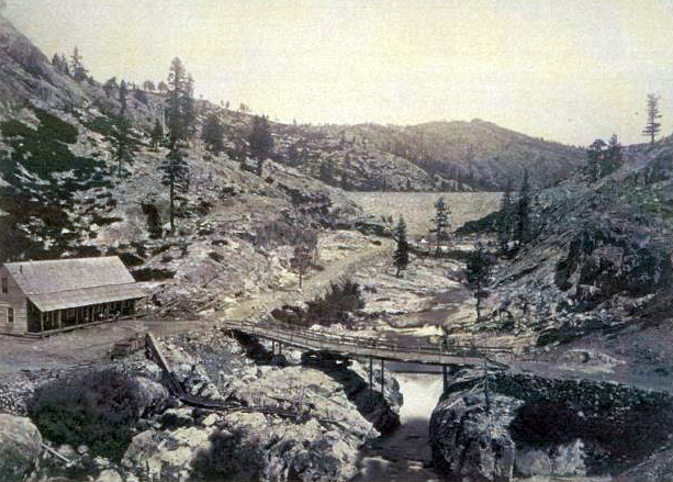 A cabin by a mountainous landscape, with a bridge over a small river. Sparse trees dot the rocky terrain.