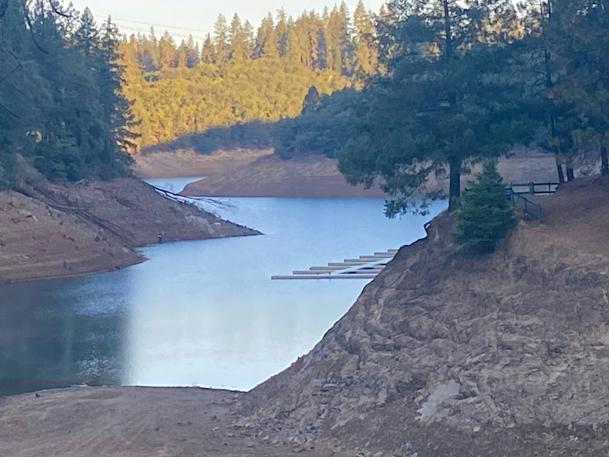 A serene reservoir surrounded by forested hills with a dock and a clear sky.