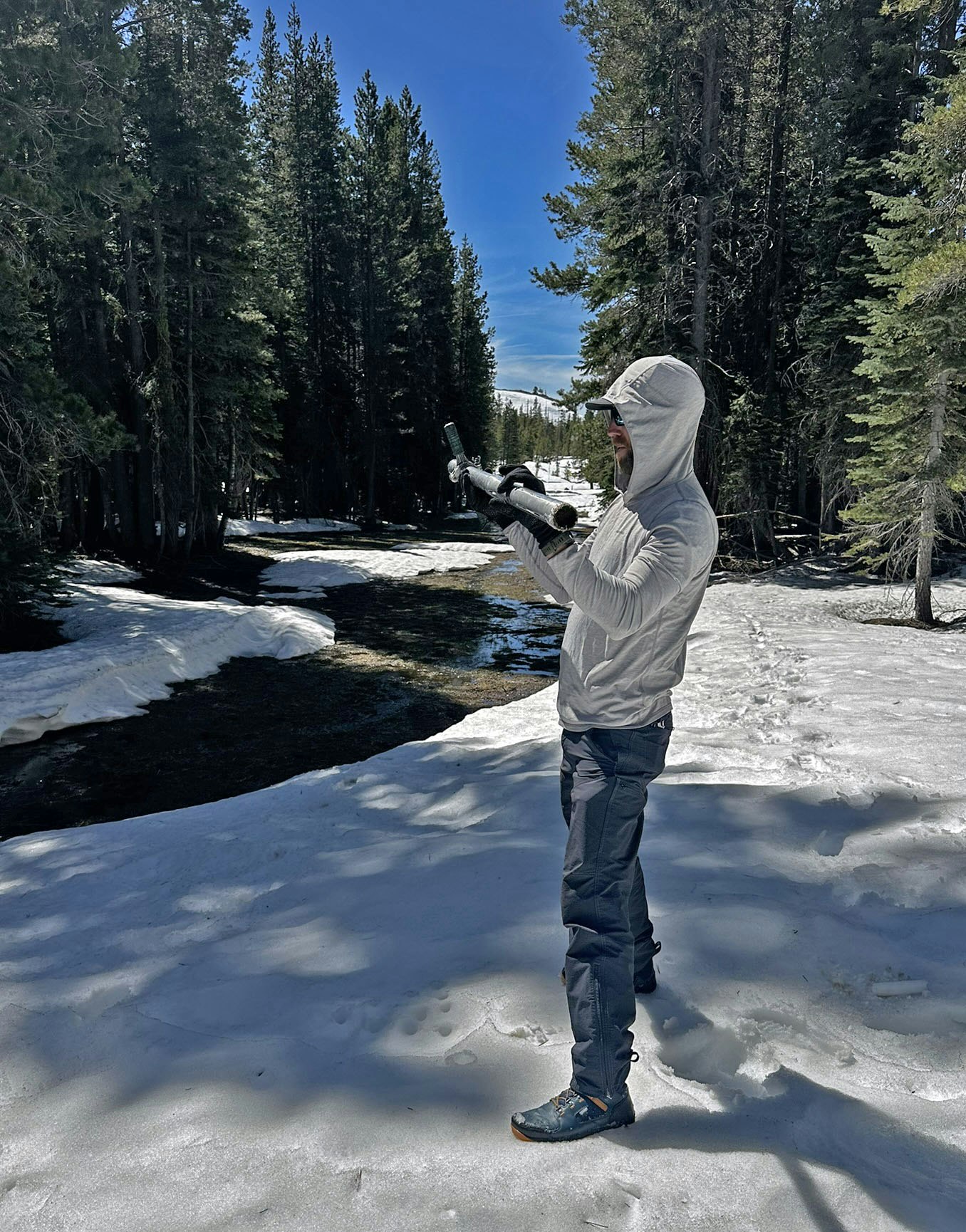 A person in a light jacket stands in a snowy forest, holding a telescope or similar device, with trees and a stream nearby.