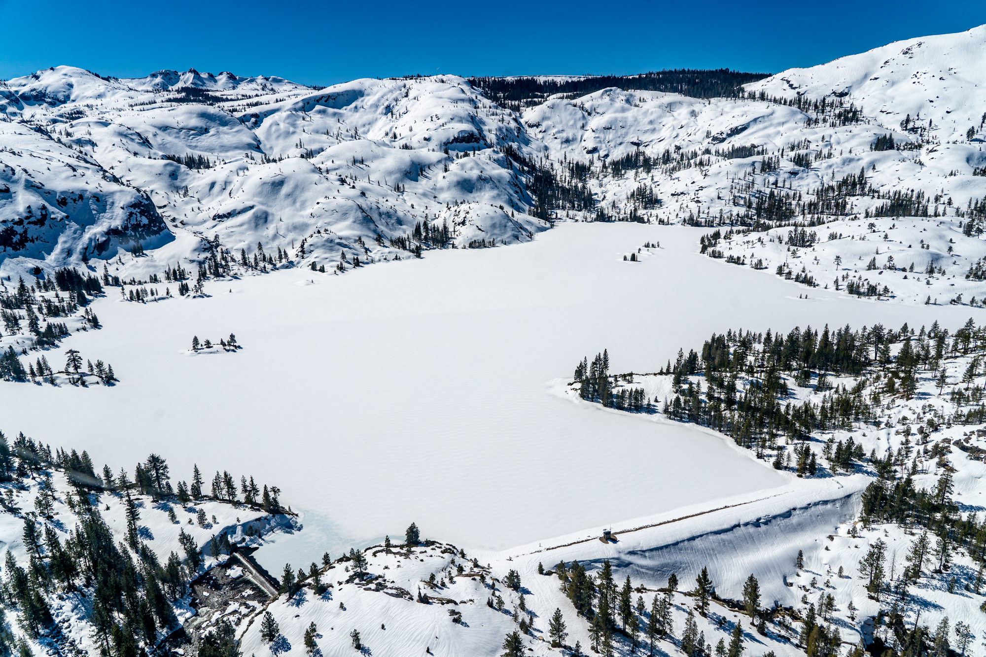 A snow-covered frozen lake surrounded by snow-dusted mountains and evergreen trees under a clear blue sky.