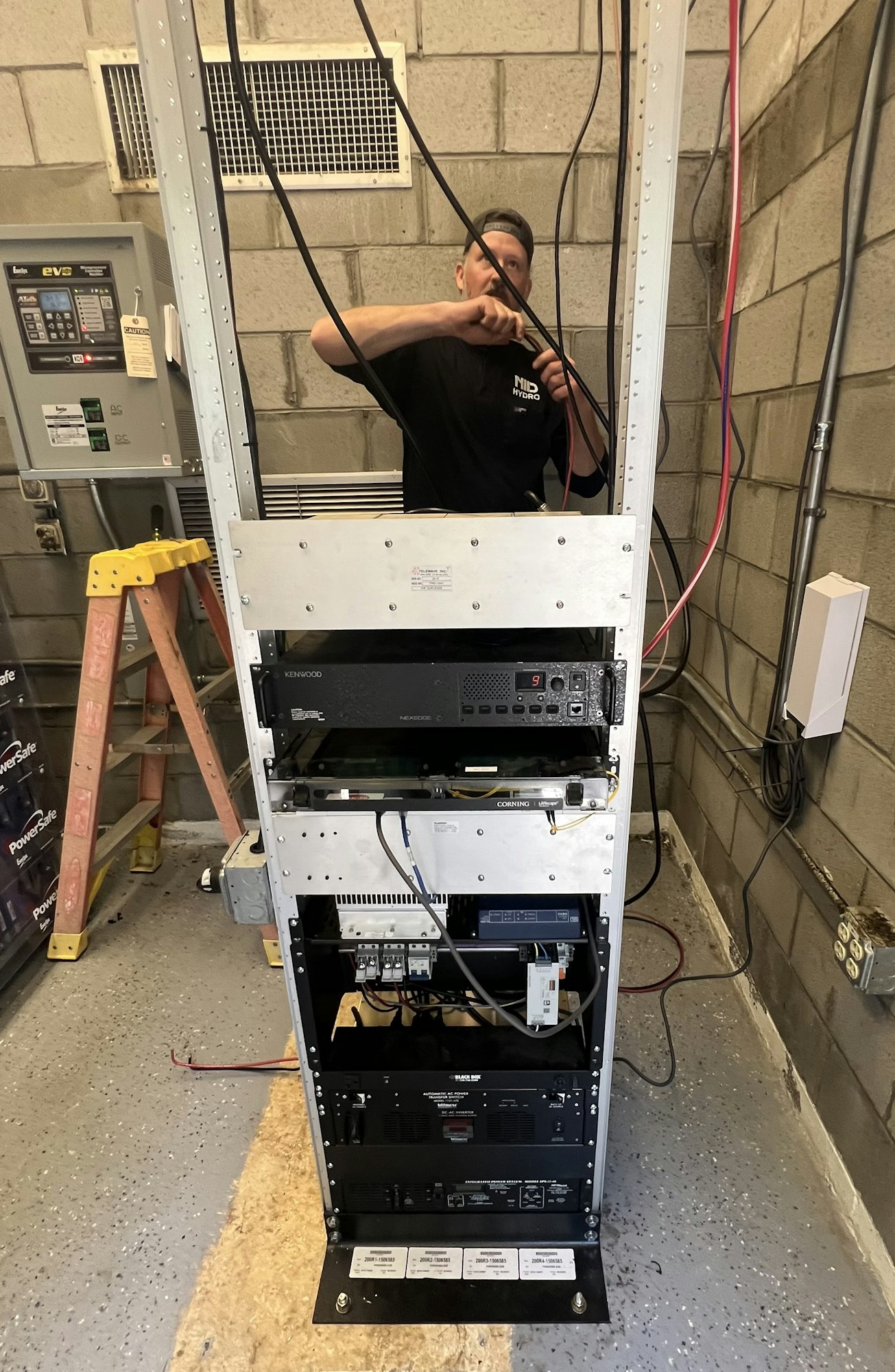 A man works on a metal rack filled with electronic equipment in a utility room, surrounded by cables and a ladder.