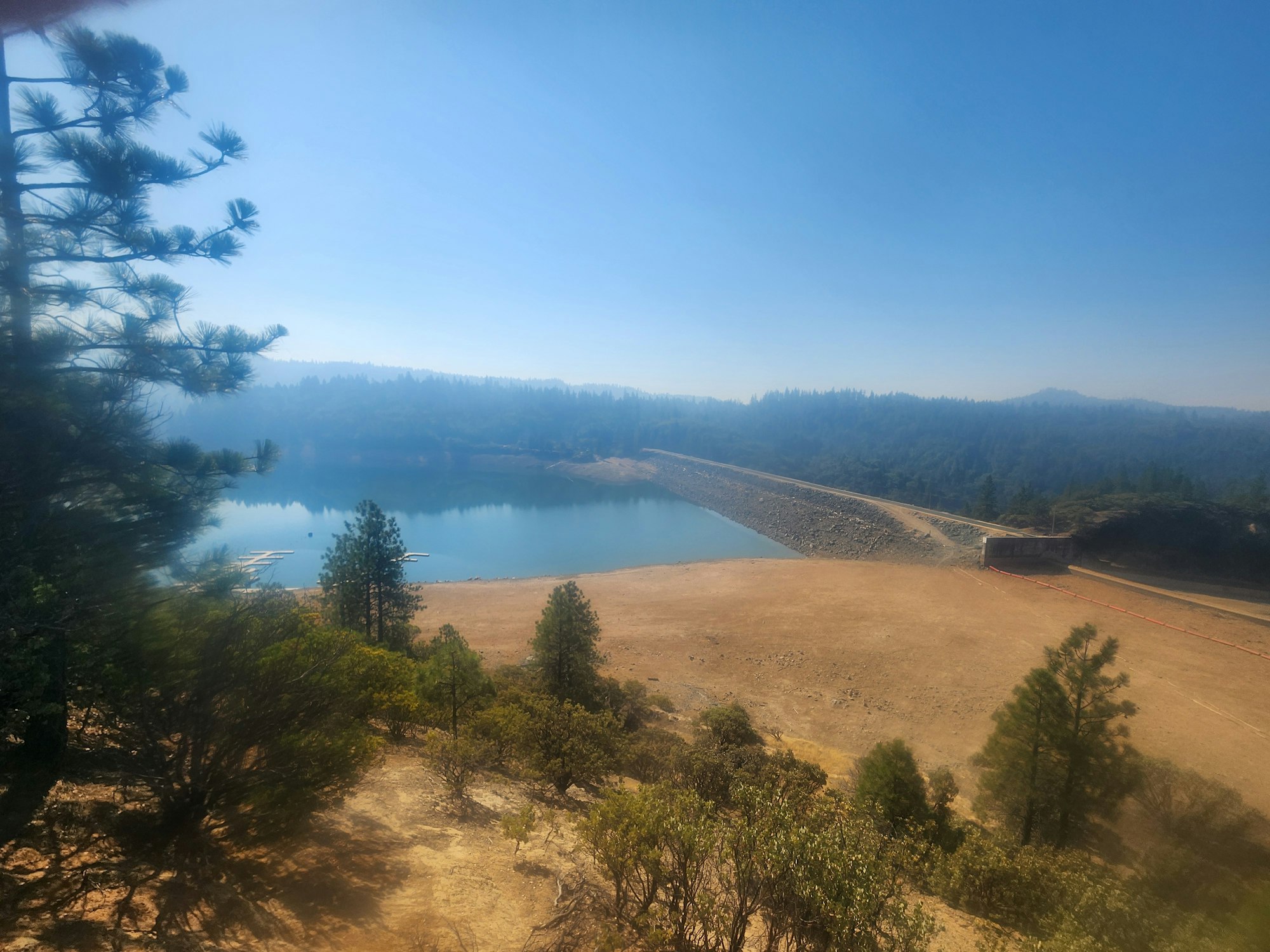 Lake with low water levels, surrounded by trees, with a dam on the right side under a hazy sky.