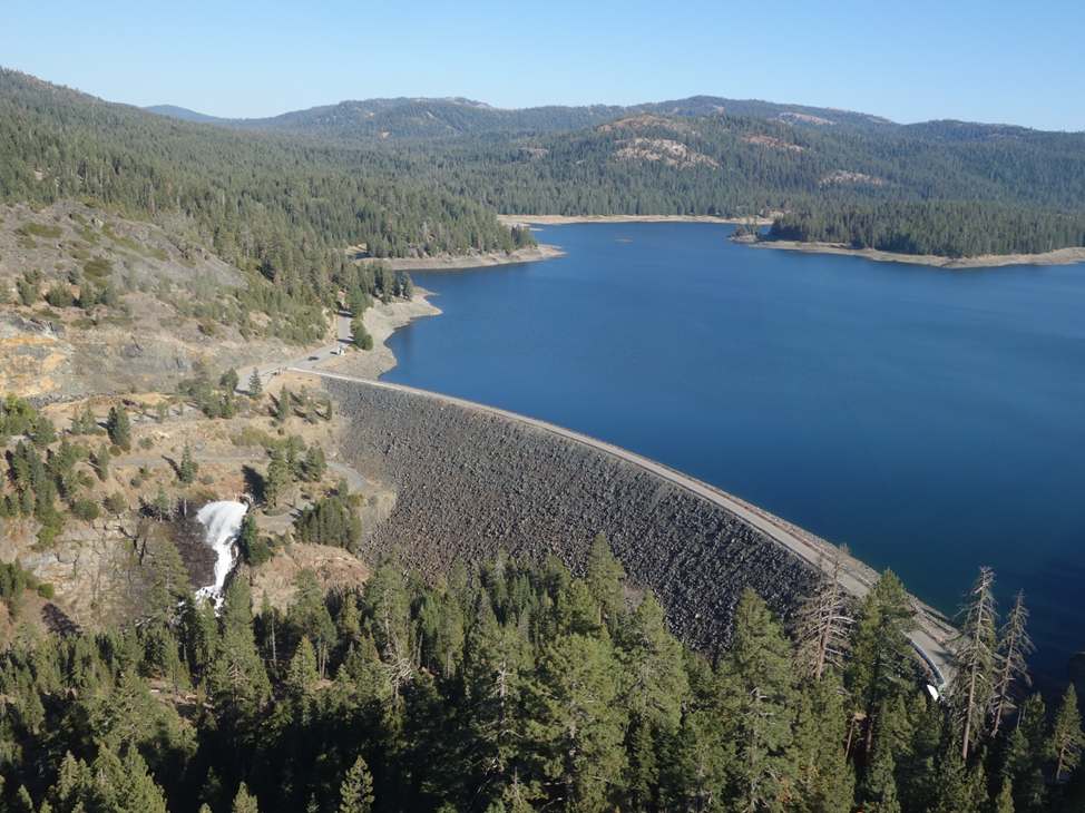 Aerial view of a lake with a dam, surrounding forests, and a small waterfall in a mountainous landscape.