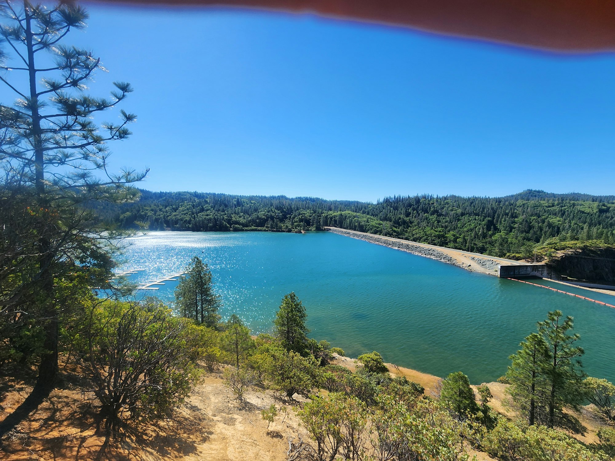 A scenic view of a blue lake surrounded by forested hills under a clear sky, with a dam on the right side.