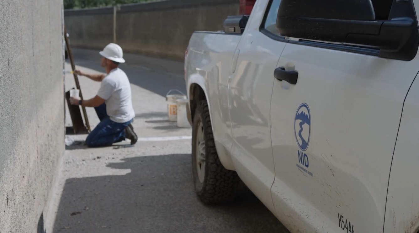 A worker in a white hat paints a wall near a white truck with a logo, suggesting maintenance or construction activity.