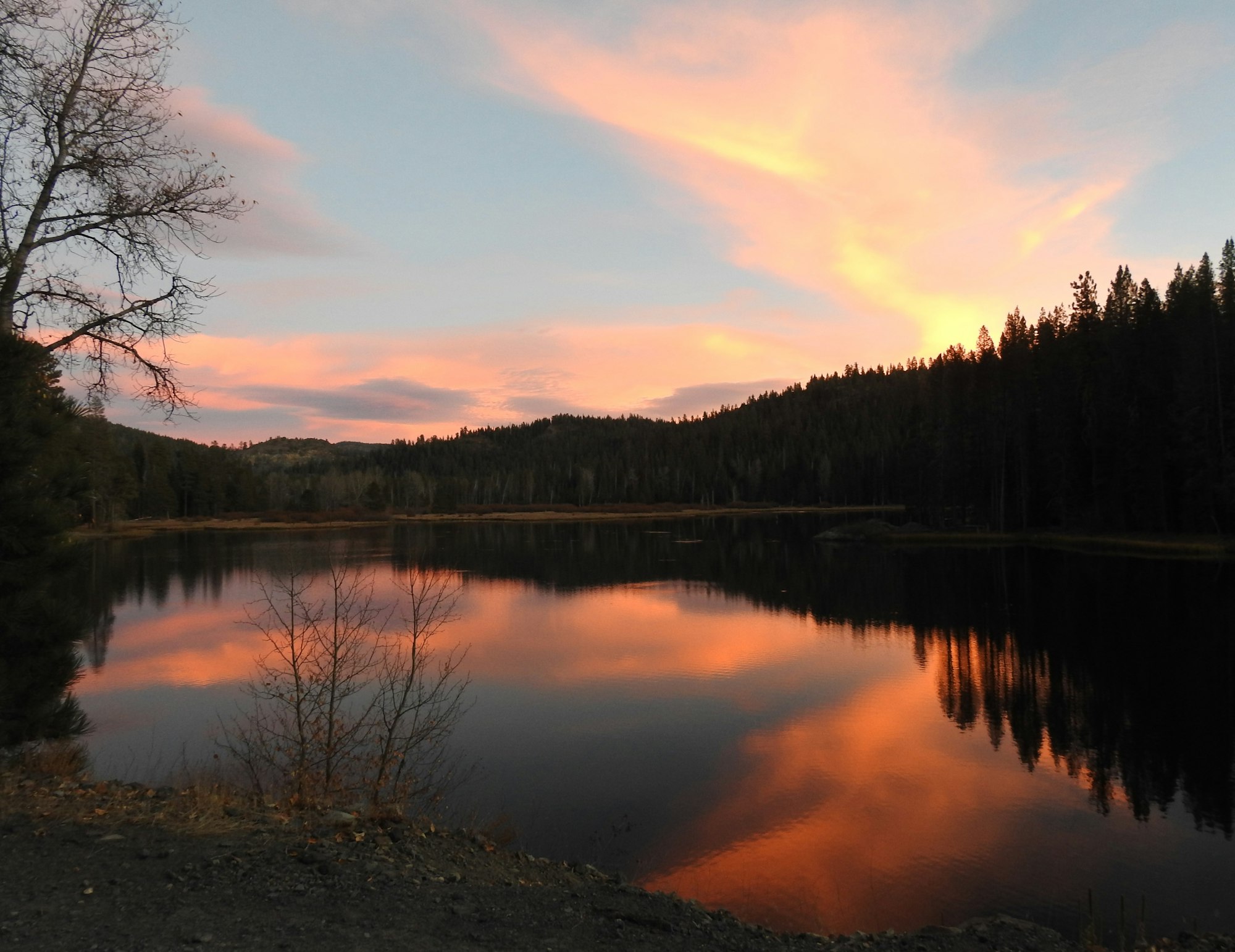 A serene lake reflecting a colorful sunset with trees and hills in the background, creating a peaceful natural scene.