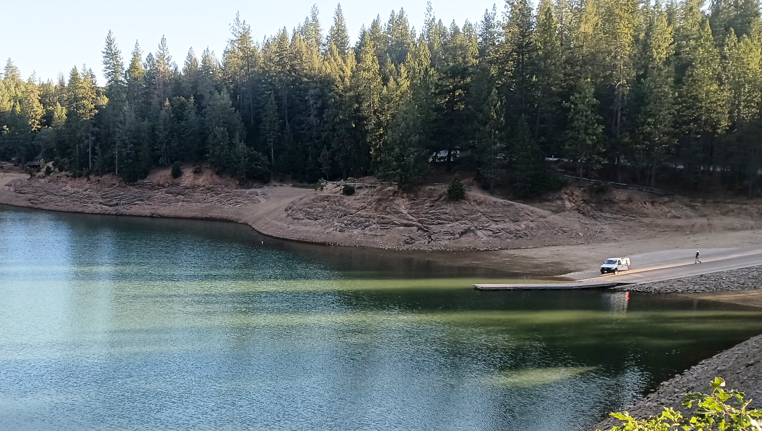 A lake with clear water, surrounded by trees and hills. A car and person on a dock by the shoreline.
