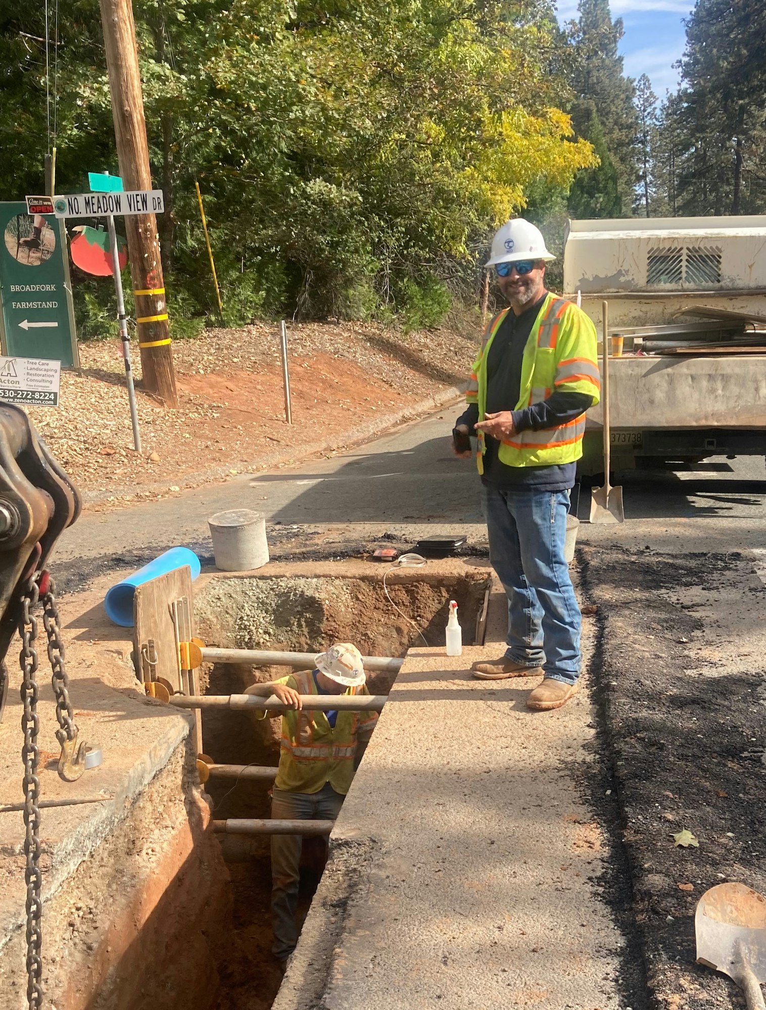 Two construction workers are digging in a trench on the side of a road, with safety gear and equipment visible.
