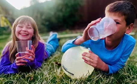 Two children are enjoying drinks and relaxing on the grass, with a soccer ball nearby, sharing smiles and fun moments.