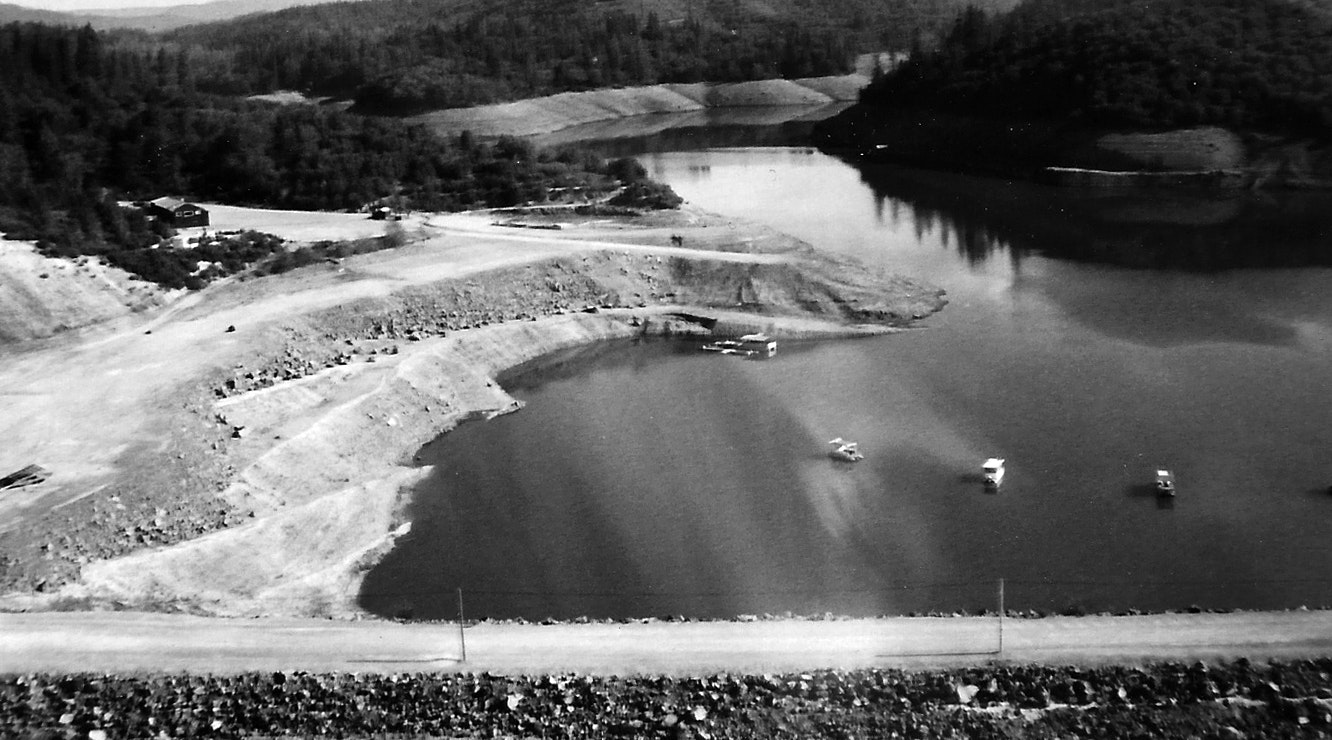 A black and white photo of a lake with boats, surrounded by forested hills and roads.