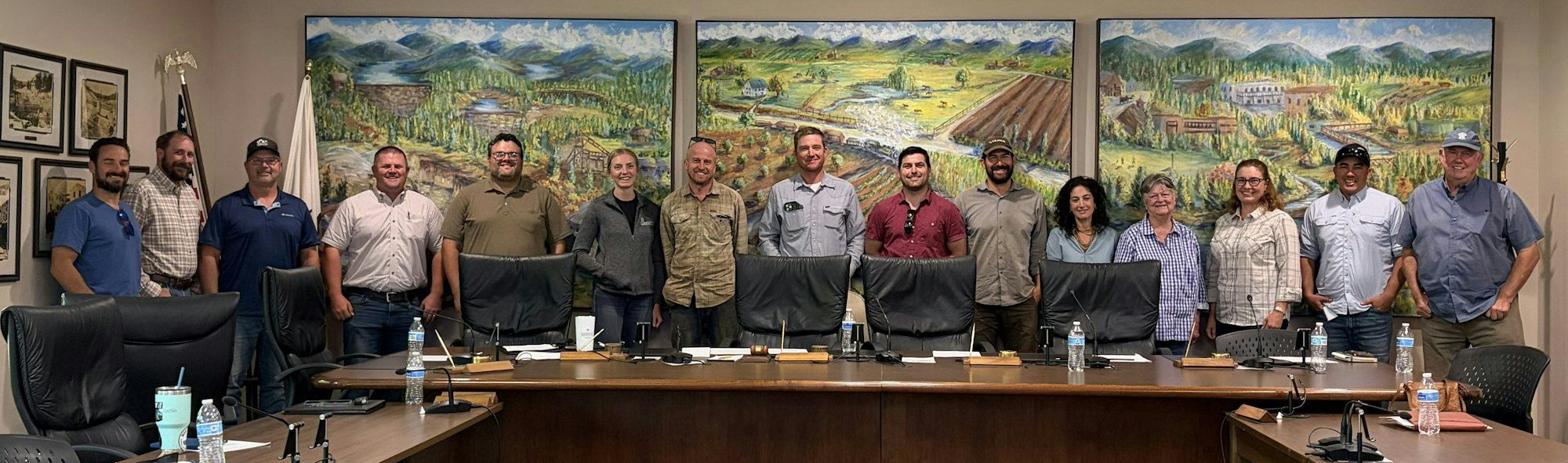 A group photo of individuals standing in front of landscape artwork, with a conference table and chairs in a meeting room setting.