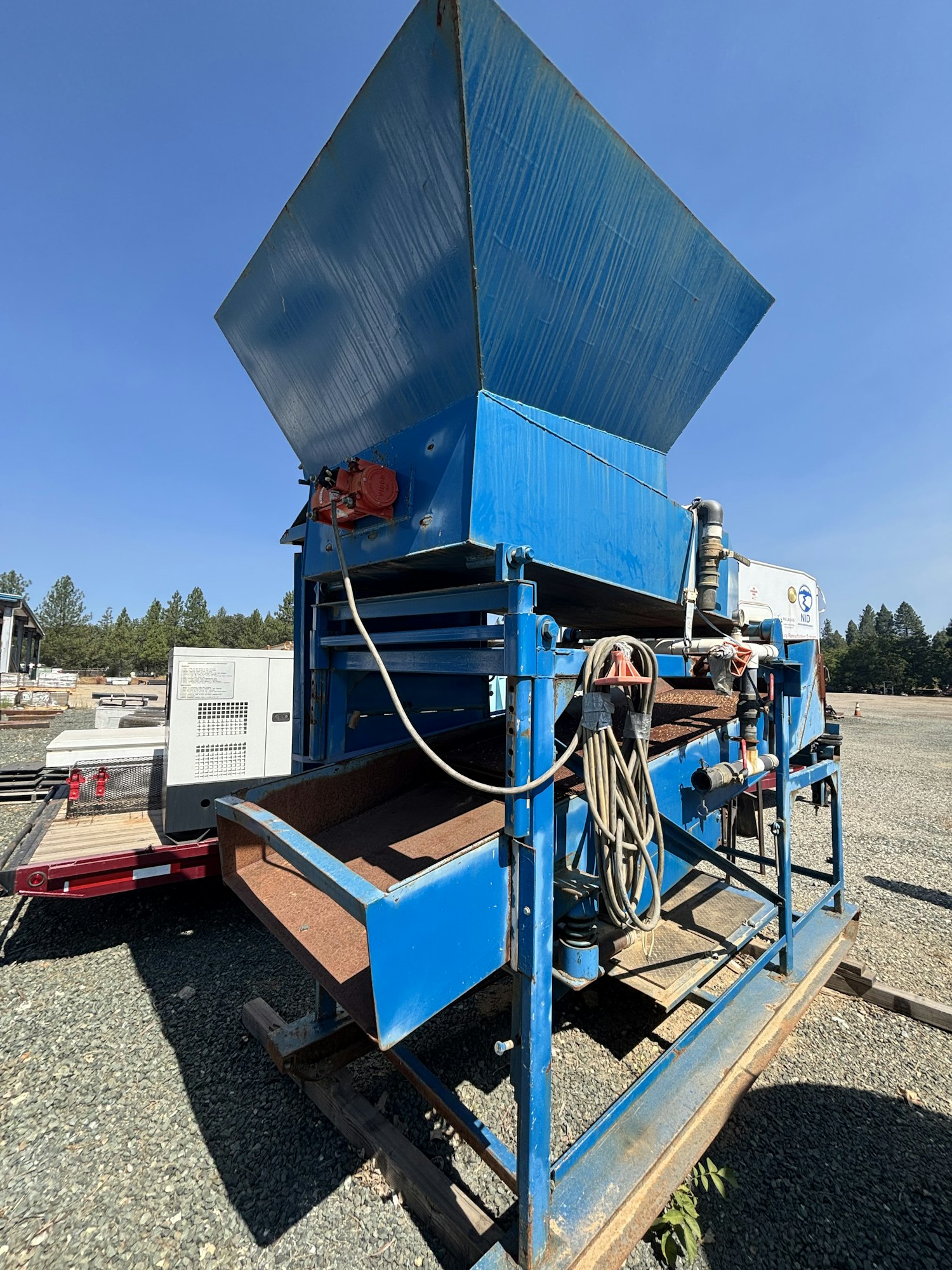 A blue industrial machine with a large hopper on top, likely used for processing materials. It's situated outdoors on gravel.