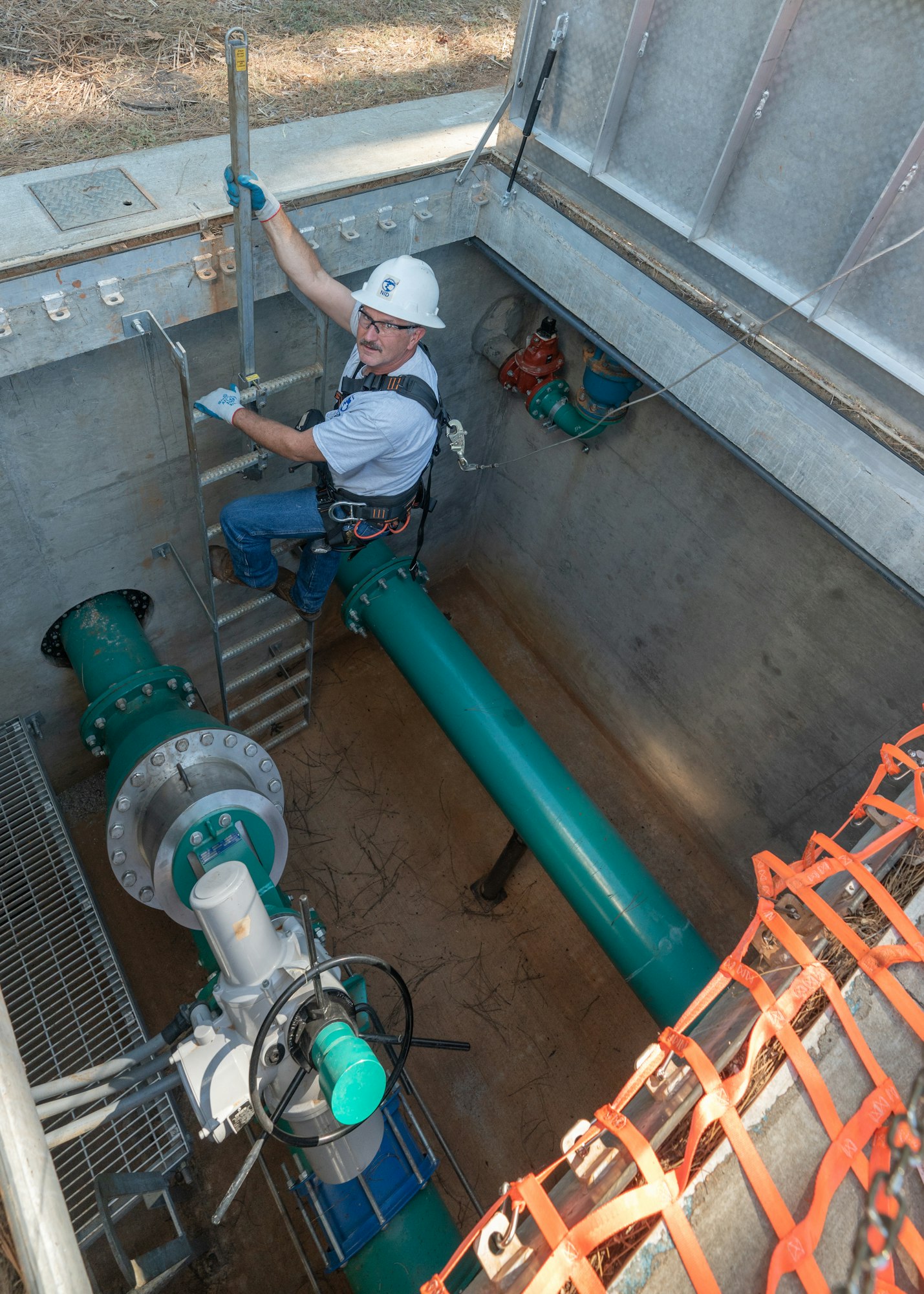 A worker in a hard hat descends a ladder into a utility area with large pipes and equipment for maintenance or inspection.