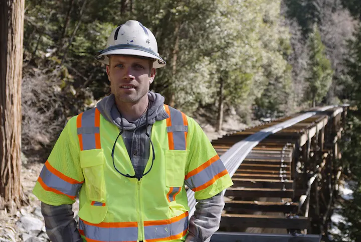 A man in a safety vest and hard hat stands near a wooden bridge in a forested area, likely indicating construction work.