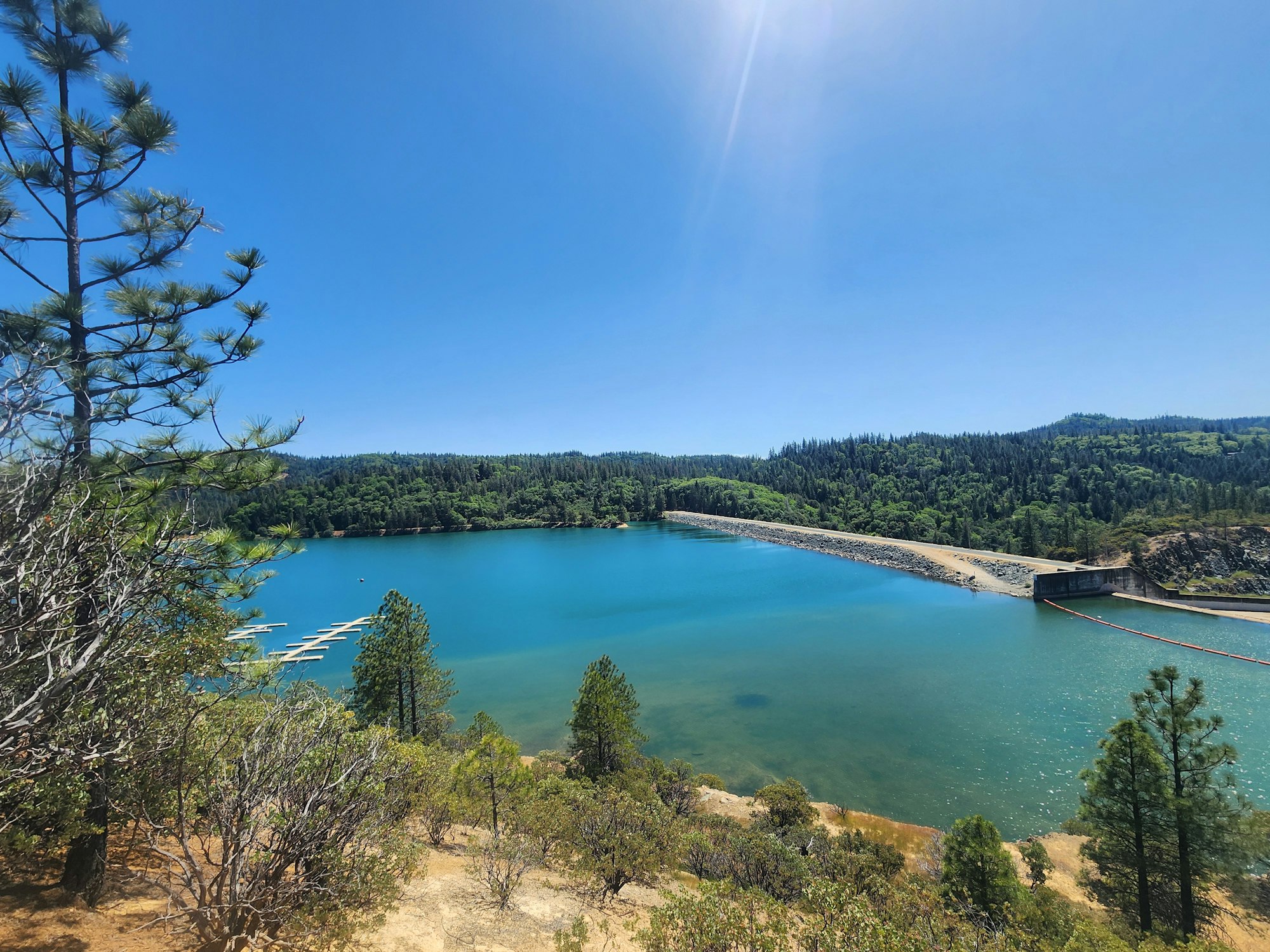 A serene lake with a dam, surrounded by forested hills under a clear blue sky.