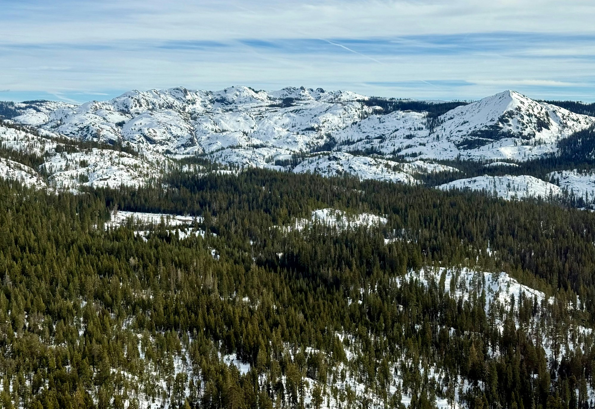 A scenic view of snow-covered mountains surrounded by dense evergreen forests under a partly cloudy sky.