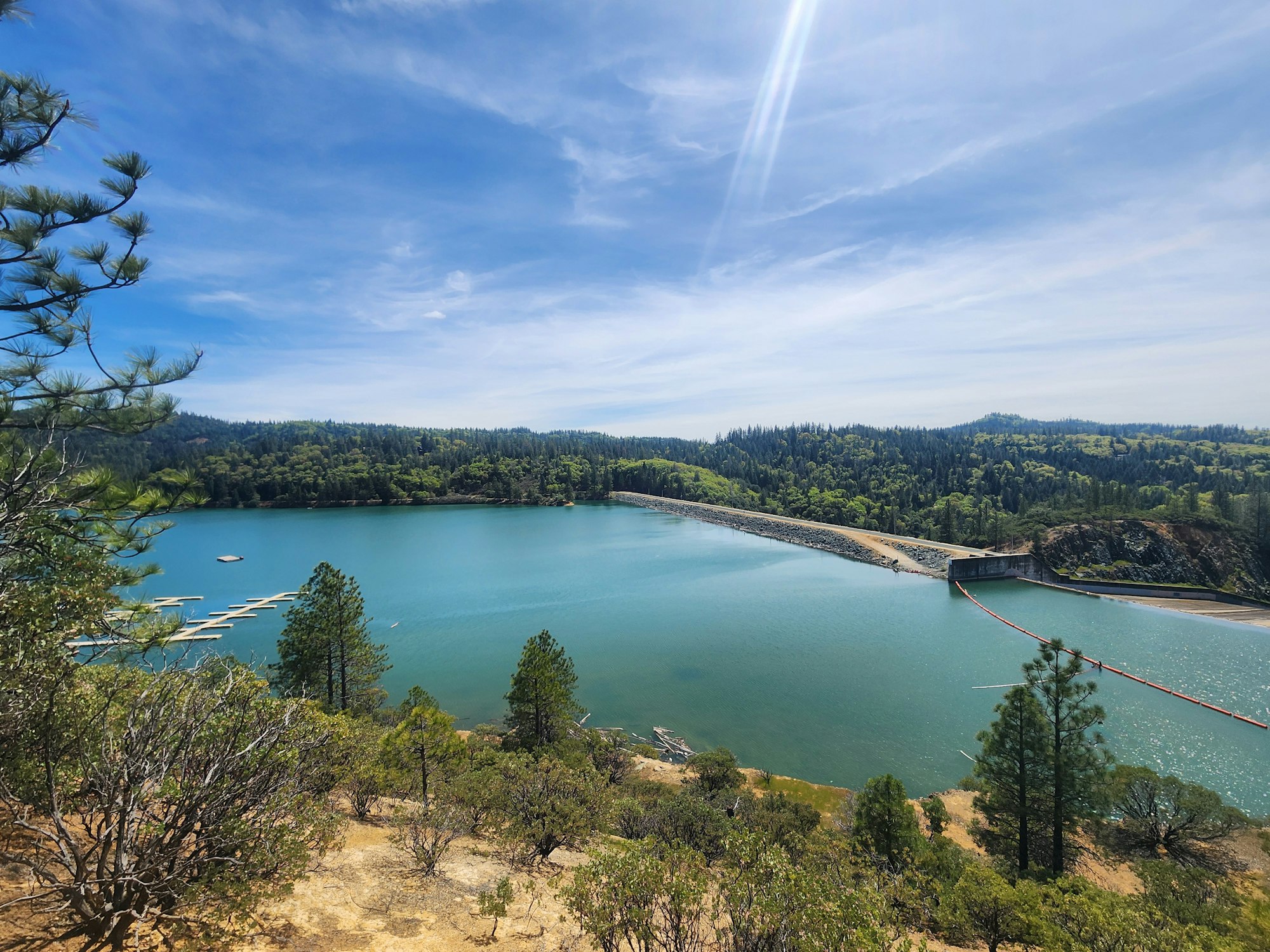 A scenic view of a lake, trees, a dam, clear skies, and a jetty with boats.