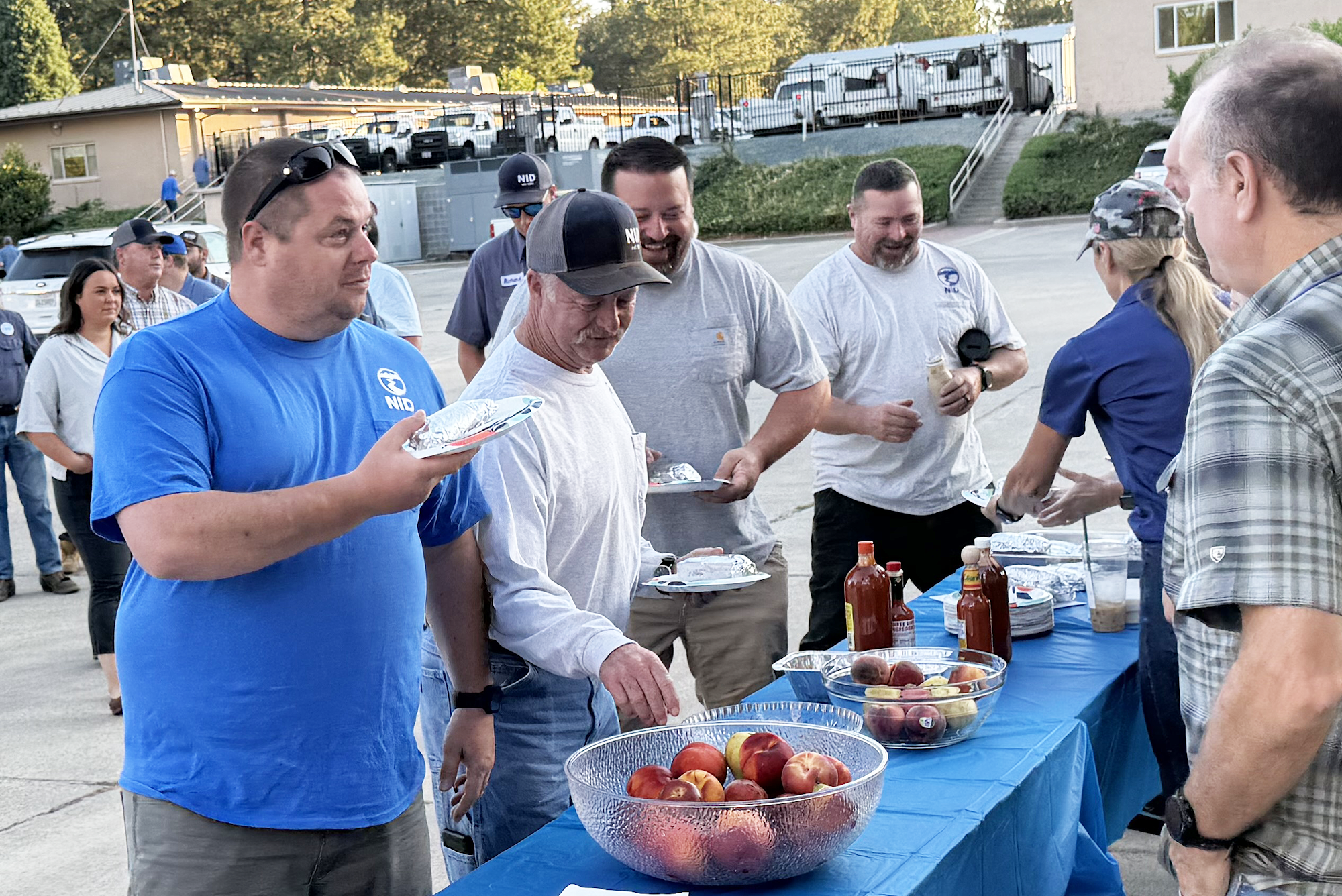 People gathered at an outdoor event, picking up food from a serving table with bowls of fruits and condiments.